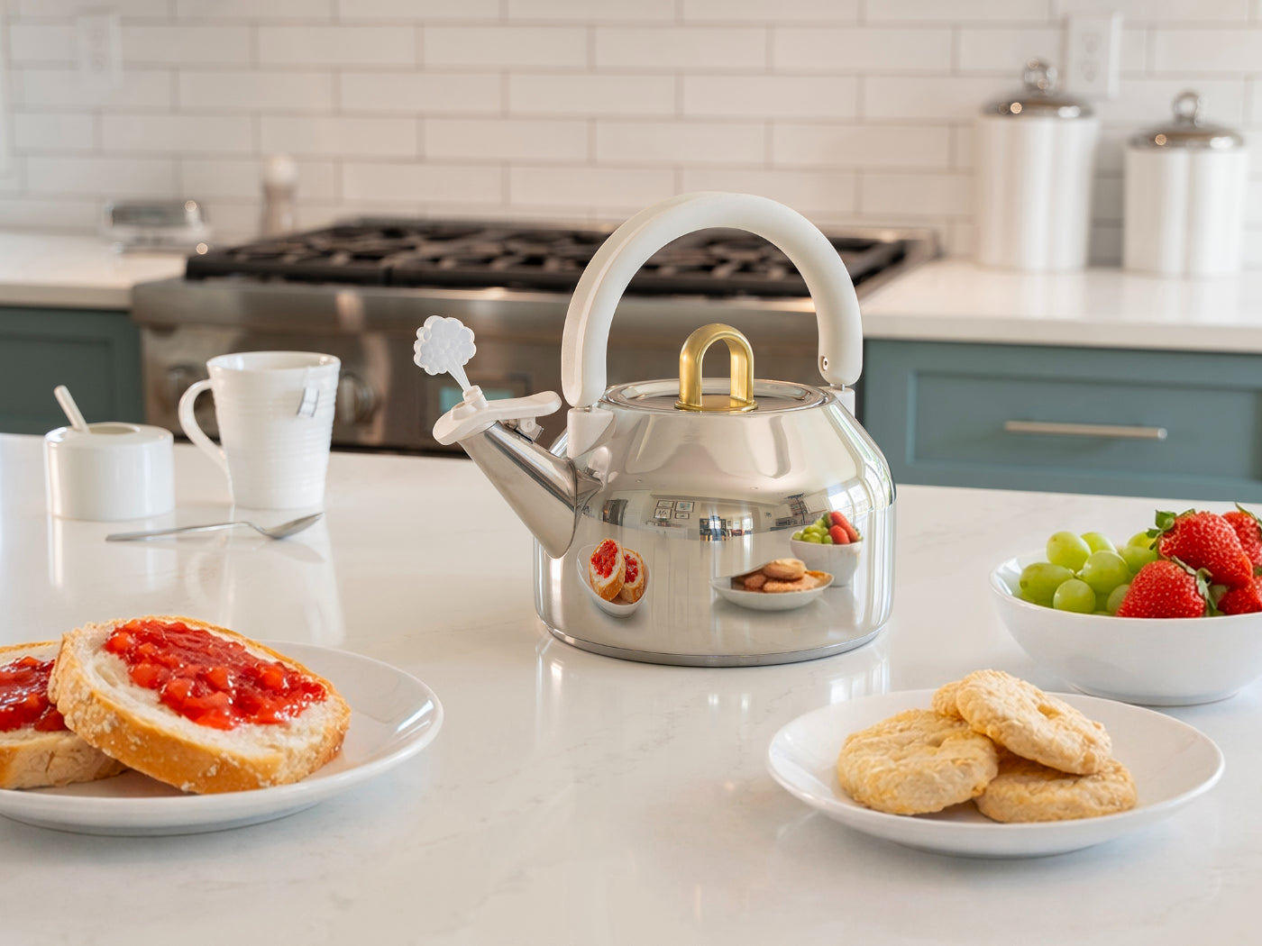 A stainless steel teapot with a white handle and lid, styled on a white kitchen countertop with breakfast food plates, a mug, and a bowl of fruit in a modern kitchen setting.