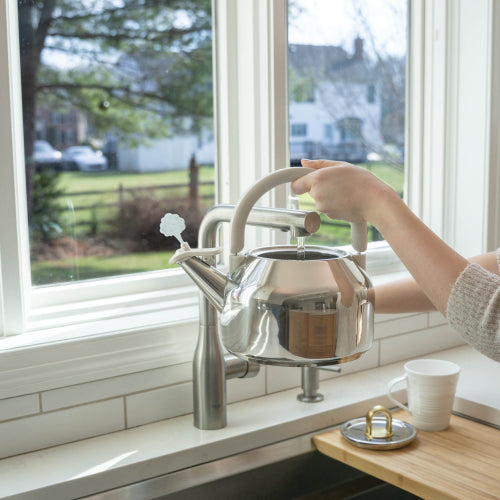A stainless steel kettle with a curved cream-colored handle is being filled with water from a modern chrome faucet at a kitchen window. A person's arm is visible holding the handle, with a white mug on a wooden tray nearby.