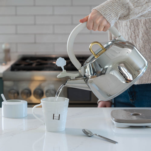 A person in a cream knit sweater pours hot water from a shiny stainless steel kettle with a white handle and gold handle accent into a white ceramic mug on a kitchen counter.