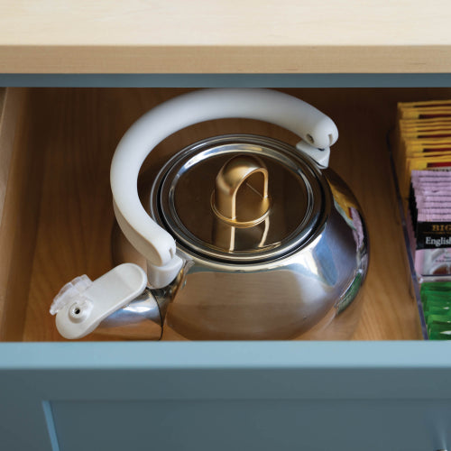 A stainless steel kettle with a white handle and lid trim, sitting inside an open wooden drawer next to tea bags.
