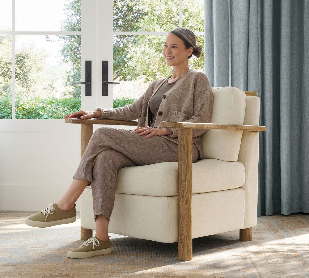 Woman sitting in a beige armchair with wooden arms in a bright room.