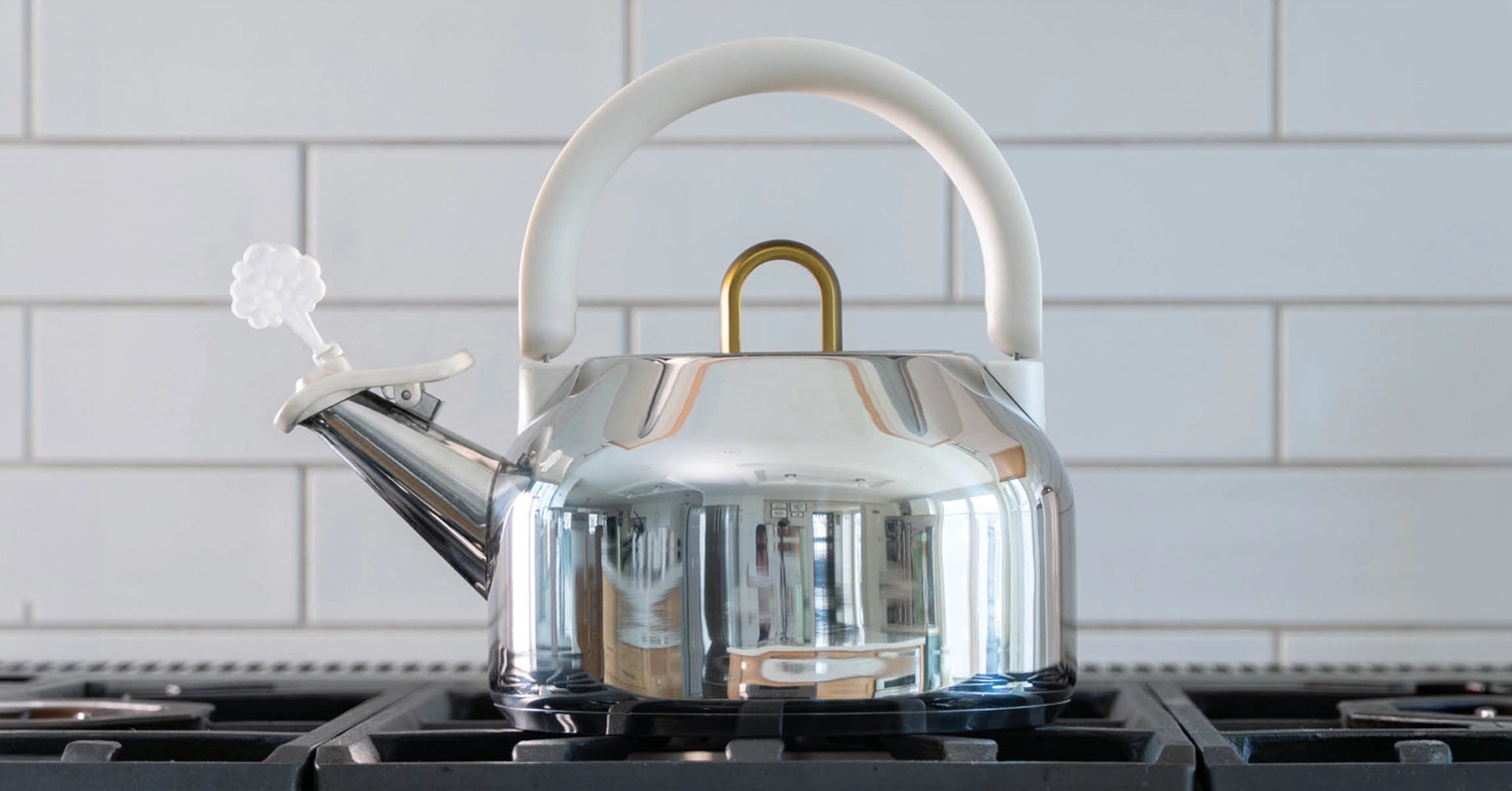 A polished stainless steel kettle with a white handle and lid, sitting on a black stovetop against a white subway tile backsplash.