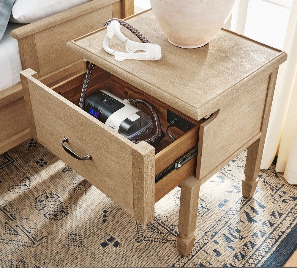 Wooden side table with open drawer containing electronic devices on a patterned rug.