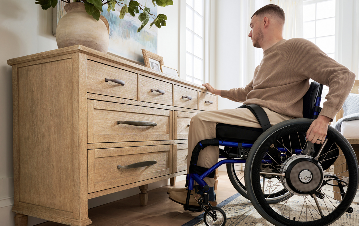 man in wheelchair holding on to edge of dresser with vase and picture frames on top of it
